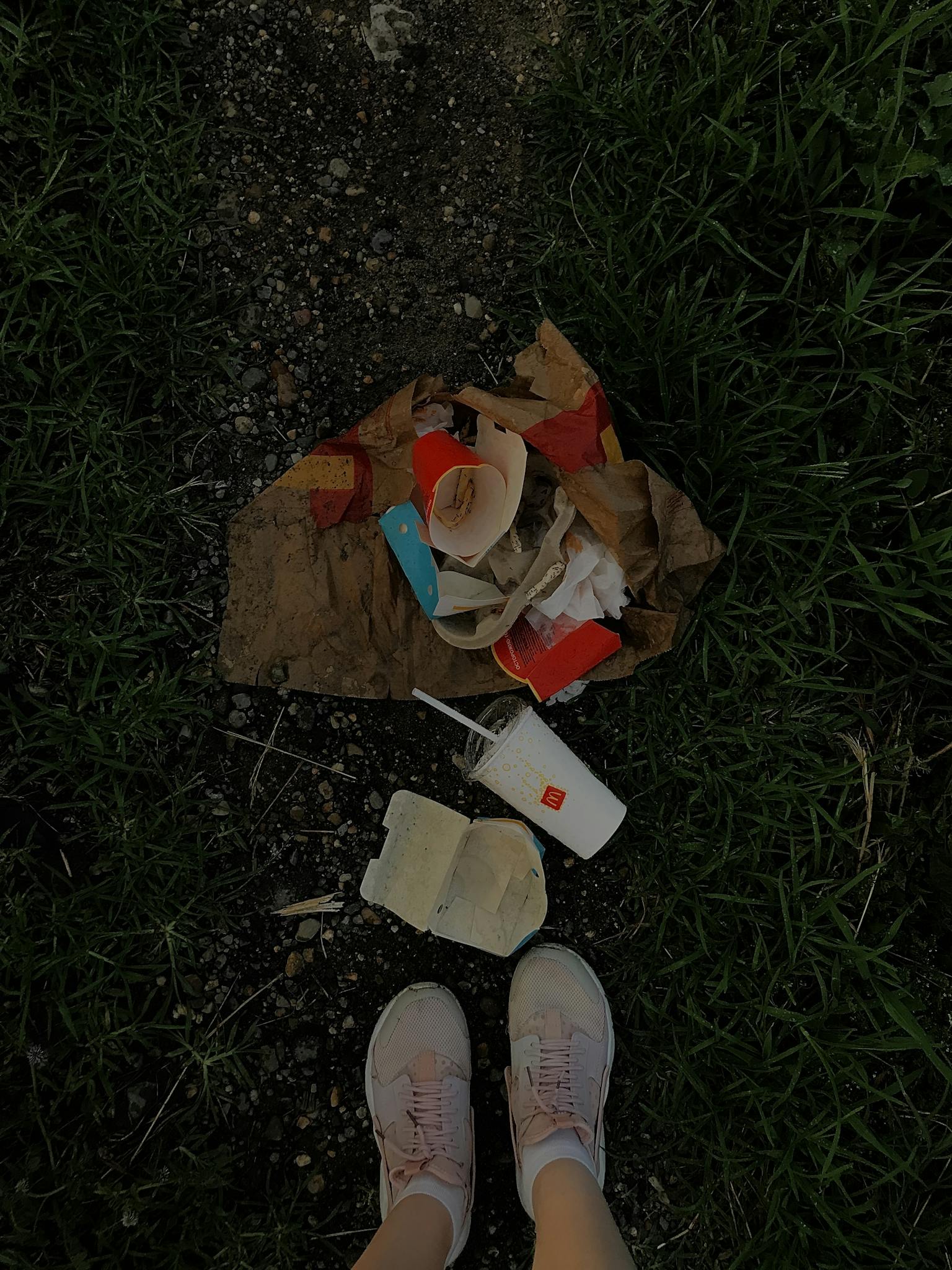 Close-up of fast food trash including cups and wrappers on a grassy path with shoes visible.